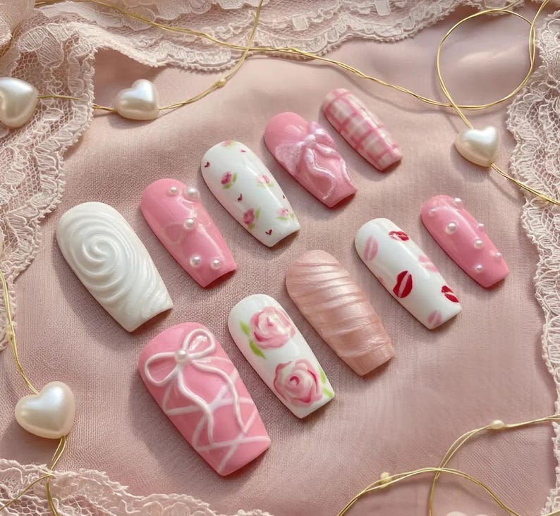 May include: A collection of artificial nails in various designs. The nails feature pink, white, and pearlescent colors with patterns like bows, roses, and lip prints. Some nails have pearl accents, and others have a textured swirl design.