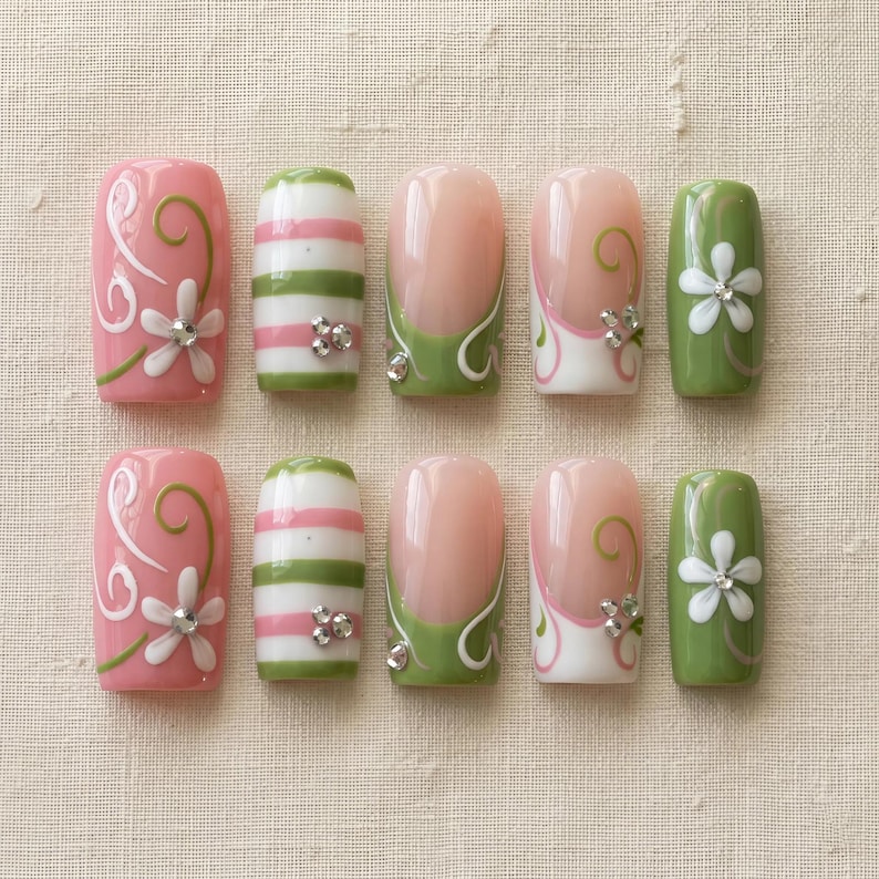 K&ouml;nnte beinhalten: Ein Set aus zehn k&uuml;nstlichen N&auml;geln in verschiedenen Designs. Die N&auml;gel zeigen rosa, gr&uuml;ne und wei&szlig;e Farben mit floralen und gestreiften Mustern. Einige N&auml;gel haben ein French-Tip-Design mit gr&uuml;nen Akzenten und Strasssteinen.