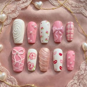 May include: A collection of ten artificial nails in various designs. The nails feature a range of pink and white colors, with embellishments like pearls, bows, floral patterns, and lip prints. The nails are arranged on a pink lace surface.