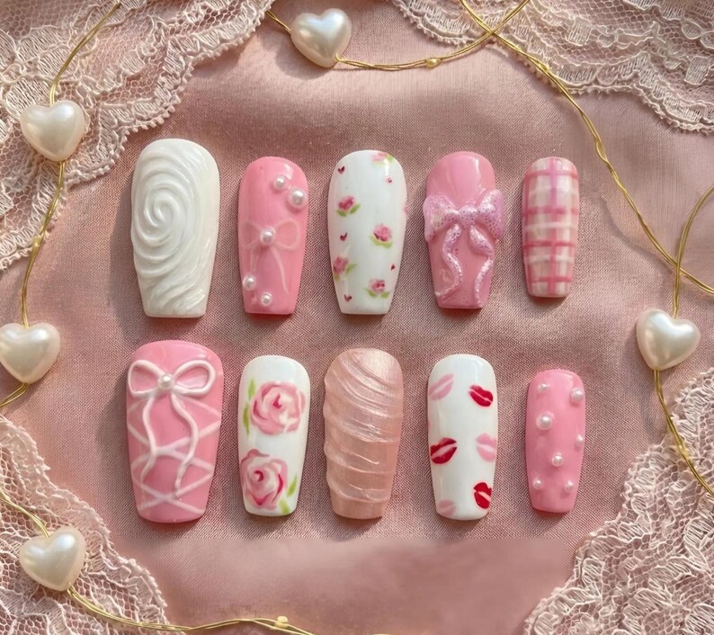 May include: A collection of ten artificial nails in various designs. The nails feature a range of pink and white colors, with embellishments like pearls, bows, floral patterns, and lip prints. The nails are arranged on a pink lace surface.