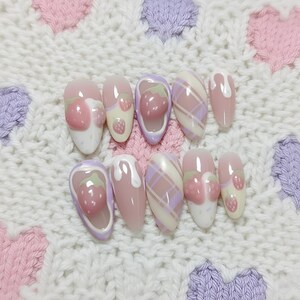May include: A set of ten artificial nails with a light pink base. Some nails feature a 3D strawberry design, while others have a plaid pattern in pink and lavender. A white, textured background with pink and purple heart shapes.