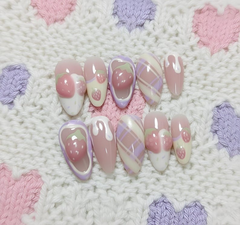May include: A set of ten artificial nails with a light pink base. Some nails feature a 3D strawberry design, while others have a plaid pattern in pink and lavender. A white, textured background with pink and purple heart shapes.