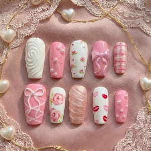 May include: A set of ten artificial nails in various designs. The nails feature a range of pink and white colors, with embellishments like bows, pearls, and floral patterns. Some nails have a textured appearance, while others showcase a plaid design.