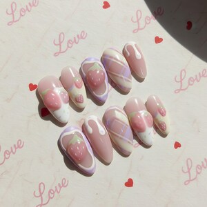 May include: A set of ten artificial nails with a light pink base. Designs include strawberries, plaid patterns, and white drizzles. The word "Love" is written in pink script on the background, along with small red hearts.