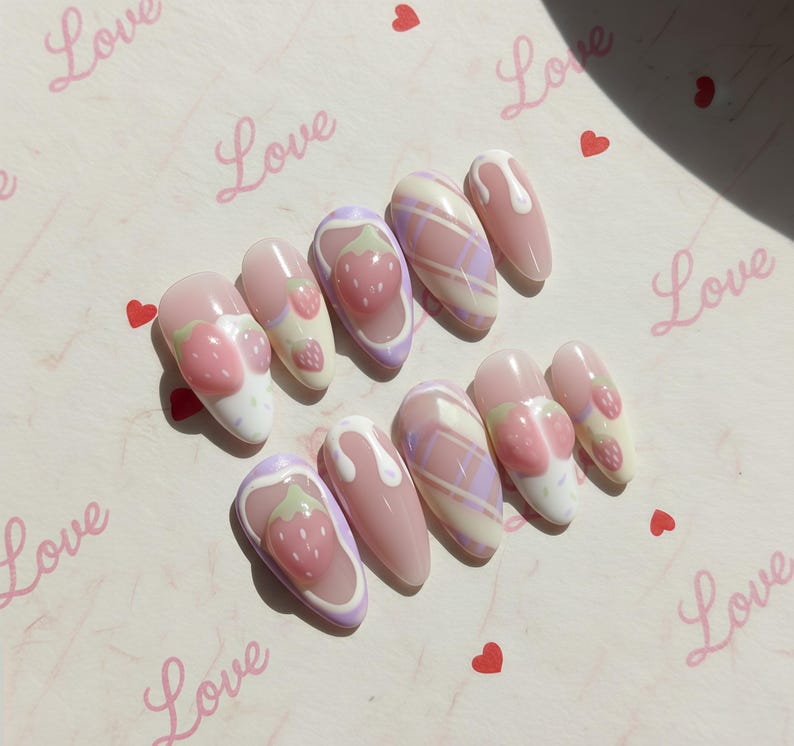 May include: A set of ten artificial nails with a light pink base. Designs include strawberries, plaid patterns, and white drizzles. The word "Love" is written in pink script on the background, along with small red hearts.