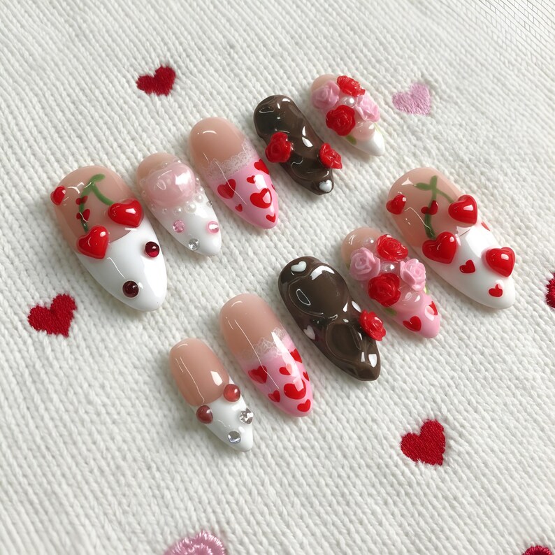 Op de afbeelding: Een verzameling kunstnagels in verschillende ontwerpen. Sommige nagels zijn beschilderd met roze en rode harten, terwijl andere rode rozen en kersen tonen. De nagels hebben een glanzende afwerking en zijn gerangschikt op een wit, getextureerd oppervlak met geborduurde rode harten.