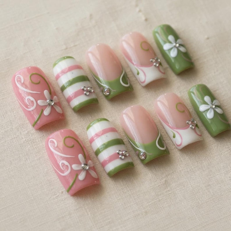 K&ouml;nnte beinhalten: Ein Set aus zehn k&uuml;nstlichen N&auml;geln in verschiedenen Designs. Einige N&auml;gel sind rosa mit wei&szlig;en Blumenakzenten, andere weisen gr&uuml;n-wei&szlig;e Streifen auf, und einige haben eine rosa Basis mit gr&uuml;n-wei&szlig;en Details. Jeder Nagel ist mit kleinen, glitzernden Verzierungen versehen.