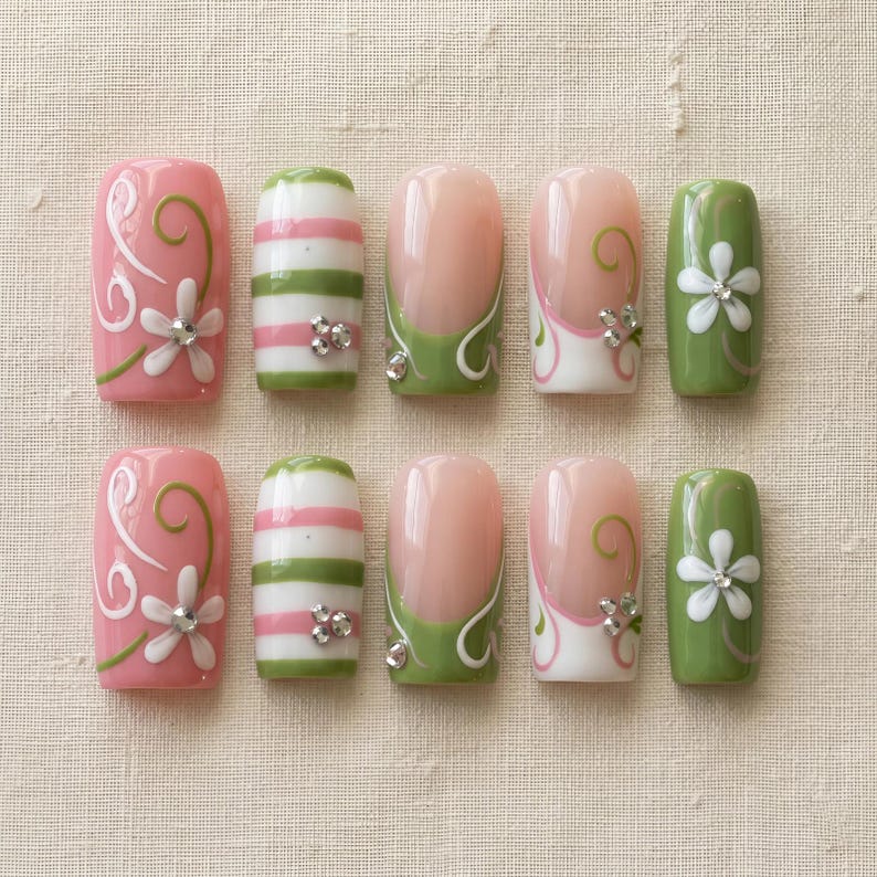 K&ouml;nnte beinhalten: Ein Set aus zehn k&uuml;nstlichen N&auml;geln in verschiedenen Designs. Die N&auml;gel zeigen rosa, gr&uuml;ne und wei&szlig;e Farben mit floralen und gestreiften Mustern. Einige N&auml;gel haben ein French-Tip-Design mit gr&uuml;nen Akzenten und Strasssteinen.