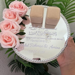 May include: A mirrored wedding ring tray with a pearl border, featuring two beige ring boxes. The tray is decorated with pink roses and includes engraved text: "And He placed between you love & mercy (Quran 30:21), Abdul Rehman & Amina 02.05.2026."