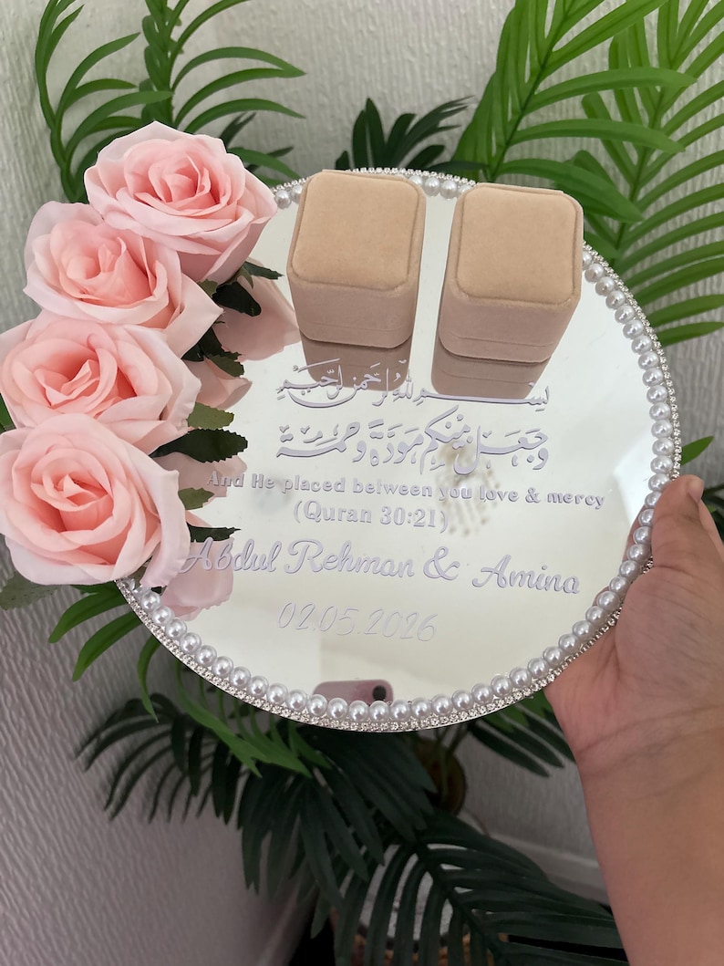 May include: A mirrored wedding ring tray with a pearl border, featuring two beige ring boxes. The tray is decorated with pink roses and includes engraved text: "And He placed between you love & mercy (Quran 30:21), Abdul Rehman & Amina 02.05.2026."