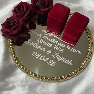 May include: A mirrored tray with a gold beaded edge, holding two red velvet ring boxes and burgundy roses. The tray has white text that reads "And we created you in pairs Quran 78:8 Sibthein & Zaynah 08.04.26". The background is white fabric.