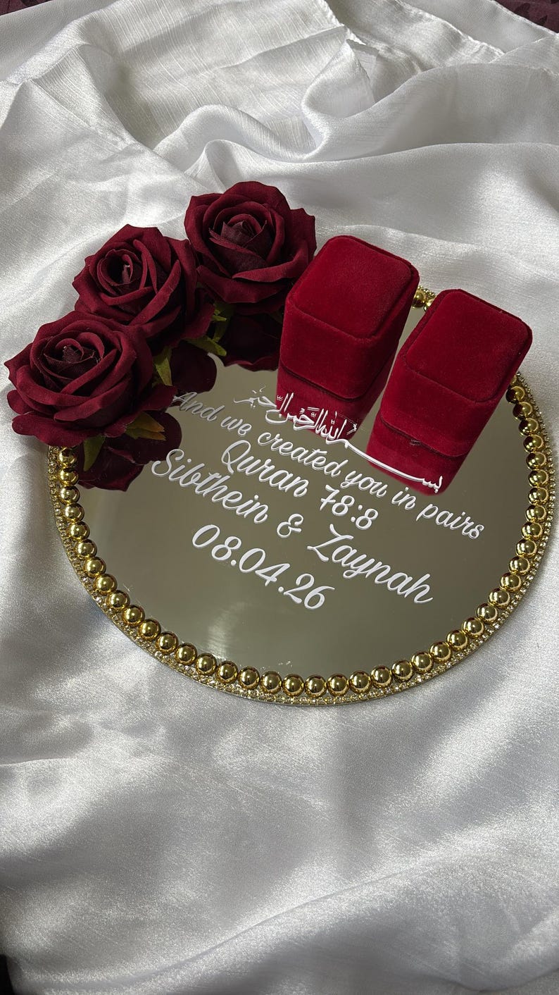 May include: A mirrored tray with a gold beaded edge, holding two red velvet ring boxes and burgundy roses. The tray has white text that reads "And we created you in pairs Quran 78:8 Sibthein & Zaynah 08.04.26". The background is white fabric.
