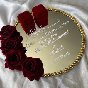 May include: A gold mirrored tray with a beaded gold border, featuring two red velvet ring boxes. The tray is decorated with red roses and inscribed with white text, including a quote and names. The date 29.11.2025 is also visible.