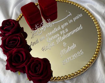 May include: A circular mirrored tray with a gold beaded border, decorated with four deep red artificial roses. Two red velvet ring boxes sit on the surface, which features white engraved text: And we created you in pairs, Quran 78:8, Hafiz Muhammad & Misbah, 29.11.2025.