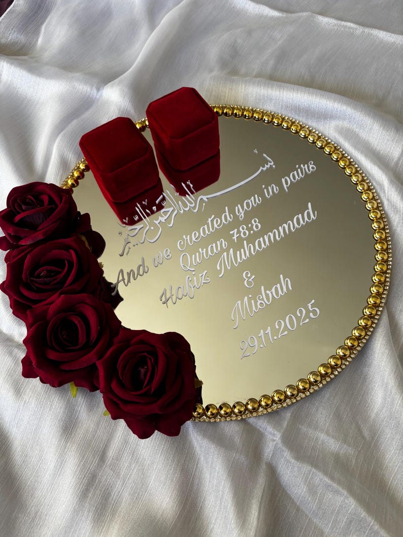May include: A gold mirrored tray with a beaded gold border, featuring two red velvet ring boxes. The tray is decorated with red roses and inscribed with white text, including a quote and names. The date 29.11.2025 is also visible.