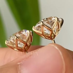 May include: A pair of gold stud earrings with a large, round, clear gemstone. The earrings have a cage-like setting around the gemstone. The earrings are held in a person's hand, with a blurred green background.