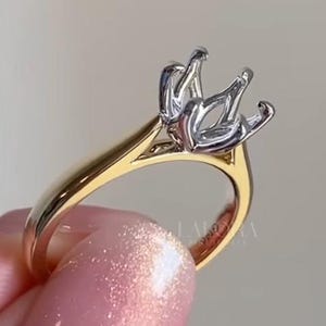 Puede incluir: Un anillo de dos tonos con una banda dorada y un engaste plateado diseñado para sostener una piedra preciosa. El anillo está sostenido por una mano. El engaste del anillo tiene cuatro garras.