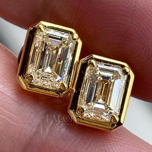 May include: A pair of gold-framed emerald-cut diamond stud earrings. The diamonds have a rectangular shape with a step-cut facet pattern. The gold setting has a polished finish.
