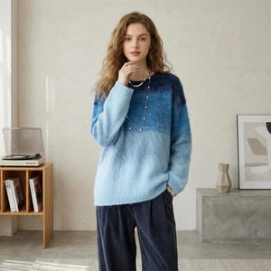 Puede incluir: Una mujer con un suéter azul degradado con cuello redondo. El suéter hace la transición de un azul marino oscuro en la parte superior a un azul claro en la parte inferior. También lleva un collar de perlas y pantalones de pana azul oscuro.