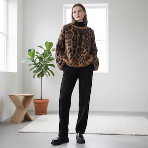 Pode incluir: Uma pessoa vestindo um suéter com estampa de leopardo marrom e preto, combinado com calças pretas e sapatos pretos. O suéter tem gola redonda e mangas compridas. As calças são de perna reta. Os sapatos são mocassins. O fundo é um quarto branco com uma janela e uma planta.