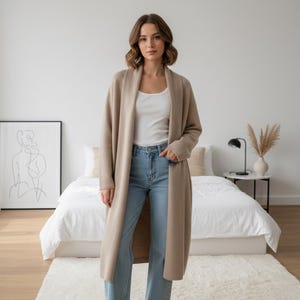 Może przedstawiać: Długi, beżowy kardigan z szalowym kołnierzem. Kardigan jest noszony na białym topie i jasnoniebieskich dżinsach. Modelka stoi w sypialni z łóżkiem, dziełem sztuki i stolikiem nocnym.