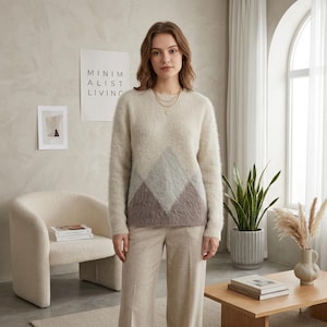 Può includere: Una donna indossa un maglione color crema con un motivo geometrico a rombi nei toni del grigio e del marrone. Indossa anche pantaloni beige abbinati. Lo sfondo include un poster minimalista con la scritta "MINIMALIST LIVING".