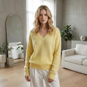 May include: A light yellow V-neck sweater with long sleeves. The sweater is paired with white pants. The model is wearing a gold necklace. The background includes a mirror, a white sofa, and plants.