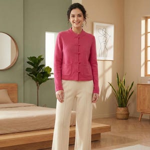 Peut inclure: Une femme porte un cardigan rose avec un col mao et des fermetures à bascule. Le cardigan a deux poches avant et est associé à un pantalon crème. La scène se déroule dans une pièce avec un lit, un miroir et une plante.