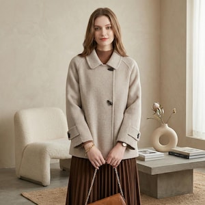 Può includere: Un cappotto di lana beige con colletto classico e due bottoni neri. Il cappotto ha maniche lunghe con polsini abbottonati. La persona indossa un dolcevita marrone e una gonna plissettata marrone. Sono visibili un braccialetto d'oro e un orologio.