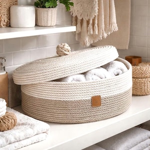 May include: A round, two-toned woven rope basket with a lid, filled with white towels. The basket is beige at the bottom and cream at the top, with a small leather tag. A decorative knot serves as a handle.