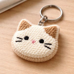 Puede incluir: Un llavero hecho a mano con forma de gato. El llavero está hecho de hilo color crema con detalles marrones en las orejas. Tiene ojos y bigotes bordados en negro y una nariz rosa. El llavero está sujeto a un aro de metal plateado.