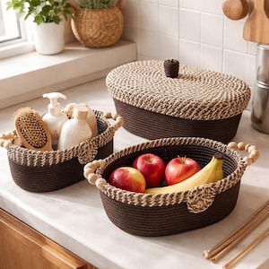May include: Three brown woven baskets with cream-colored trim and handles. One basket holds apples and a banana, another holds toiletries, and the third has a lid. The baskets are on a white countertop.