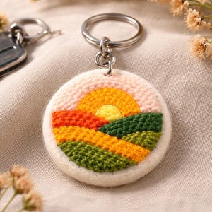 May include: A circular keyring with a stitched landscape design. The design shows a yellow sun setting over rolling hills in orange, green, and yellow. The keyring is off-white with a silver metal ring attached.