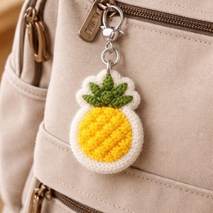 May include: A handmade pineapple keychain. The keychain features a yellow pineapple with green leaves, outlined in white. It is attached to a silver-colored metal chain and clasp. The background is a beige backpack.