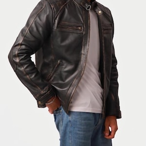 Puede incluir: Chaqueta de cuero marrón oscuro con diseño acolchado en los hombros y cuello alzado. La chaqueta tiene dos bolsillos en el pecho con cremallera y cierre frontal con cremallera. La chaqueta se lleva con una camiseta blanca y vaqueros azules.