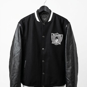 Puede incluir: Chaqueta universitaria negra con mangas de cuero y un parche con el logo de los Raiders. La chaqueta tiene una parte delantera abotonada, puños a rayas blancas y negras y un cuello blanco. El logo de los Raiders es un escudo plateado y negro con un pirata.