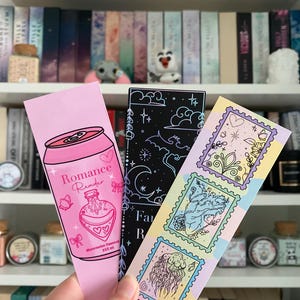 Könnte beinhalten: Drei dekorative Lesezeichen mit einzigartigen Designs. Ein pinkfarbenes Lesezeichen zeigt ein Limonaden-Dosen-Design mit dem Text "Romance Reader". Ein schwarzes Lesezeichen hat ein himmlisches Design. Das dritte Lesezeichen hat drei Briefmarken-Designs.
