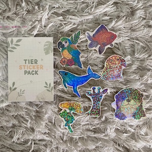 Könnte beinhalten: Eine Sammlung farbenfroher Tieraufkleber, darunter ein Papagei, ein Goldfisch, ein Wal und ein Otter, zusammen mit einer "TIER STICKER PACK" Karte. Die Aufkleber haben einen holografischen Effekt und sind auf einer flauschigen, grauen Oberfläche angeordnet.
