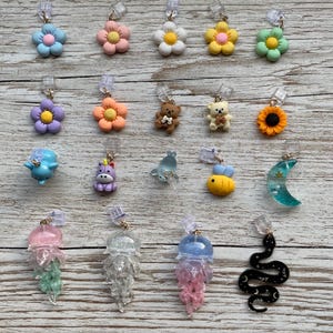 Könnte beinhalten: Eine Sammlung von neunzehn kleinen Anhängern in verschiedenen Formen, darunter Blumen, Teddybären, ein Einhorn, eine Biene, eine Mondsichel, Quallen und eine Schlange. Jeder Anhänger ist an einer durchsichtigen Kunststoffschlaufe befestigt.