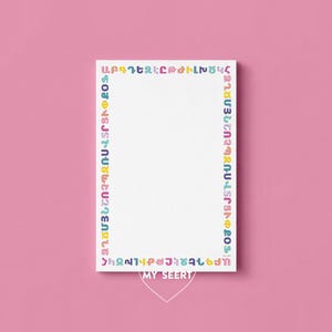 May include: A white notepad with a colorful Armenian alphabet border. The letters are in various colors, including pink, blue, yellow, and green. The notepad is on a pink background. The text "MY SEERT" is at the bottom.