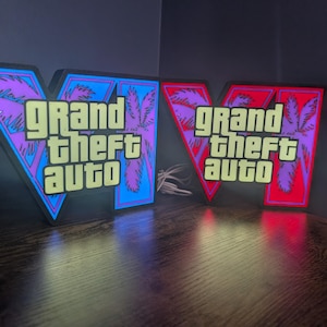 May include: Two V-shaped illuminated signs featuring the text Grand Theft Auto in yellow block letters. The left sign displays a blue background with purple palm tree silhouettes, while the right sign features a red background with purple palm trees.