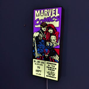 May include: A rectangular, illuminated wall sign featuring a Marvel Comics graphic with characters in purple, red, and blue tones. The sign displays the text Marvel Comics, pricing of $1.00 US and $1.25 CAN, the date 70 SEPT, and the Comics Code Authority seal.