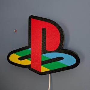 May include: A colorful PlayStation logo light fixture. The logo features a red 'P' shape, a blue, green, and yellow design, and a black outline. A white power cord extends from the bottom. The fixture is mounted on a gray wall.