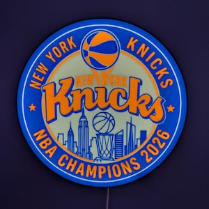 May include: A circular LED wall sign featuring the New York Knicks logo in blue and orange. The design includes a basketball, the team name, a New York City skyline silhouette, a championship trophy, and the text New York Knicks NBA Champions 2026.