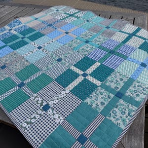 May include: A square patchwork quilt featuring a grid pattern of various blue, teal, and white fabrics. The design incorporates alternating blocks of solid colors, floral prints, gingham checks, and thin stripes, all stitched together with visible quilting lines.