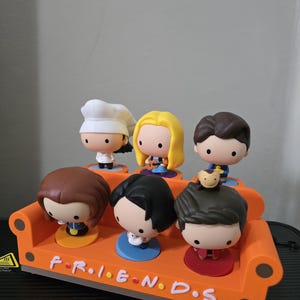 May include: A collection of six Friends character figures, each with a distinctive hairstyle and outfit, arranged on an orange sofa-shaped stand. The stand features the word "FRIENDS" in white lettering. The figures have a cartoon aesthetic.