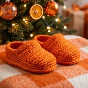 May include: A pair of bright orange crochet slippers featuring a thick, textured yarn construction with a rounded toe and a low-cut ankle opening, designed with a solid color pattern and a dense, hand-stitched appearance throughout the exterior.