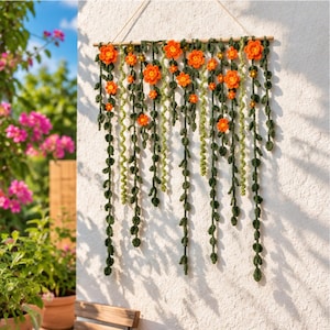 Puede incluir: Un tapiz de pared que presenta múltiples hilos de enredaderas de color verde oscuro tejidas a ganchillo y flores de color naranja brillante suspendidas de un listón de madera horizontal con follaje de varias longitudes.