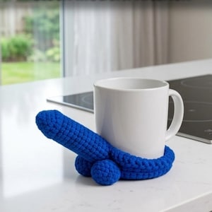 Può includere: Una tazza in ceramica bianca è posizionata sopra un sottobicchiere all'uncinetto blu brillante. Il sottobicchiere presenta un'estensione di forma fallica che sporge dalla base, realizzata con un motivo a maglia testurizzato e annodato.