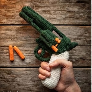 May include: A crocheted toy revolver in dark green with a white handle and orange accents on the cylinder, accompanied by two small orange crocheted cylindrical projectiles. The item features a textured yarn construction throughout the entire design.